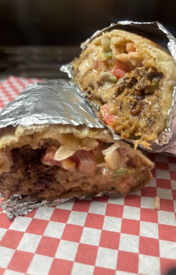 Las dos mitades de un burrito envuelto en papel aluminio, abiertas para mostrar carne, tomate y cebolla picados en una salsa cremosa, sobre papel a cuadros rojos y blancos